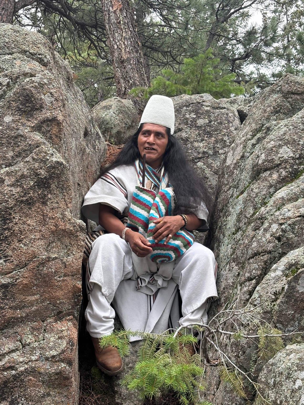 Mamo Lorenzo, an Arhuaco elder in traditional white dress and conical hat, seated among the granite boulders and pines at Blue Sky Sanctuary