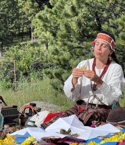 Maná, Janpi Awaq, in a beaded headband and white dress, seated in prayer among the pines with a ceremonial offering laid before her