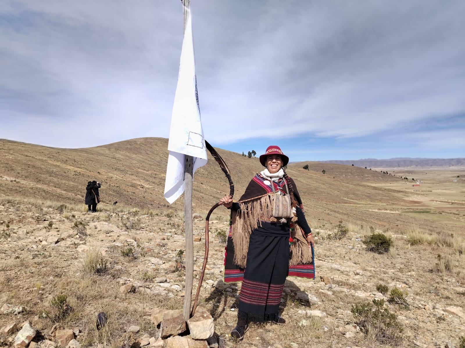 Maná in traditional Andean dress and red hat, smiling beside a white ceremonial banner in the high open landscape of the Andes